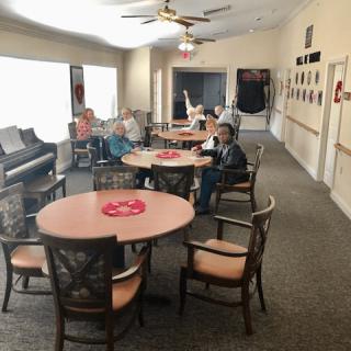 A community room with tables and chairs, occupied by people, likely in a senior living facility.