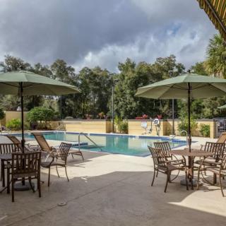 A serene outdoor pool area with tables, chairs, and umbrellas surrounded by trees under a cloudy sky.