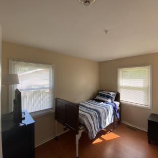 A simple bedroom with a single bed and two windows, furnished with a dresser and a lamp.