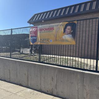 A promotional banner displayed on a fence in front of a building under clear blue skies.
