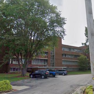 A multi-story building with a parking lot, surrounded by trees on an overcast day.