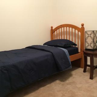 A simple minimalist bedroom with a single wooden bed, dark bedding, and a nightstand with a lamp.