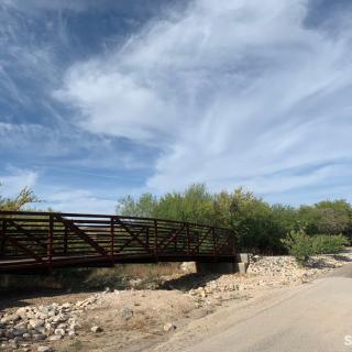 A metal bridge extends across a dry, rocky area under a vast blue sky with scattered clouds.