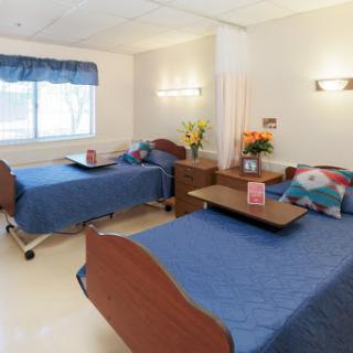 A well-lit shared patient room in a hospital with two beds, each equipped with blue bedding and bedside furniture.