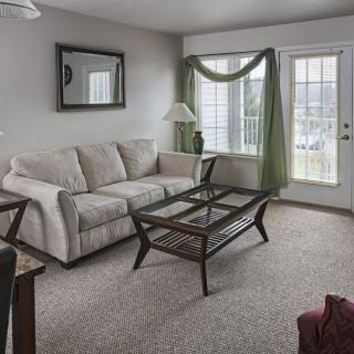 A cozy living room with a beige sofa, glass-top coffee table, and green curtains framing the window.
