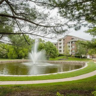 A scenic view of a pond with a fountain, surrounded by greenery and a residential building.