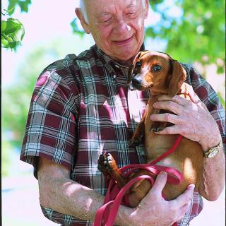 An elderly man holding a small dog outdoors on a sunny day.