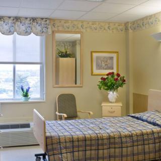 A hospital room with a single bed, flowers on a nightstand, and a chair by the window.