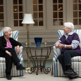 Two elderly women engage in a conversation while sitting indoors.