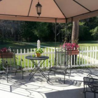 A cozy outdoor patio area with a gazebo, featuring a table and chairs surrounded by flowers and a white picket fence.