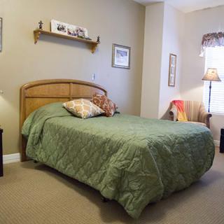 A cozy bedroom featuring a bed with a green quilt, wall art, and a standing lamp.