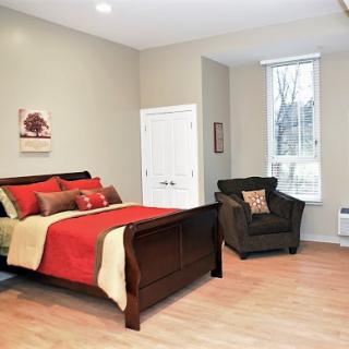 A cozy bedroom featuring a wooden bed with red and beige bedding, a comfortable armchair, and natural light streaming through the window.