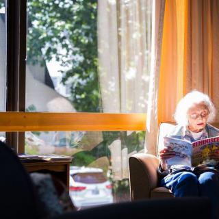 An elderly person reads a newspaper in a sunlit room by the window.