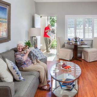 An elderly woman sits on a sofa with her pet dog, as a visitor enters the bright and cozy living room.