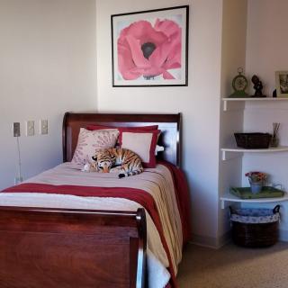 A cozy bedroom featuring a single bed with red and beige bedding, adorned with a stuffed animal. A large floral painting hangs above the bed, and built-in shelves are neatly arranged along the wall.