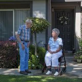 An elderly couple spending time together outside their house, with one person sitting in a wheelchair.