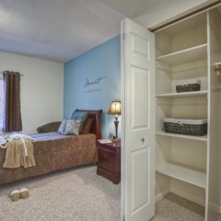 A cozy bedroom featuring a bed, nightstand with a lamp, and an open wardrobe with shelves.