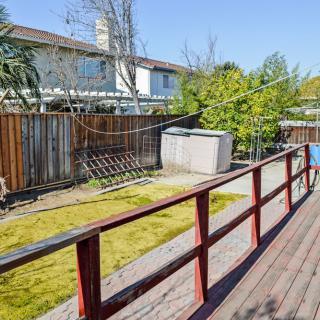 A sunny backyard with a wooden deck, green lawn, and trees along the fence.