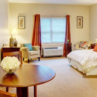 A cozy bedroom with warm lighting, featuring a bed, armchair, and a small table near a window with curtains.