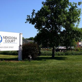 A sign for Nekoda Court stands in a grassy area with a tree and building in the background.
