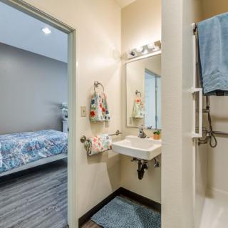 A modern bathroom with a view into an adjacent bedroom, featuring a sink, mirror, and shower area.