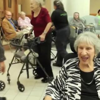 A group of elderly people socializing in a nursing home setting, with some using wheelchairs and walkers.
