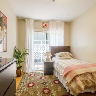A cozy bedroom featuring a single bed, decorative rug, and a potted plant beside a window with curtains.