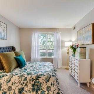 A cozy and well-decorated bedroom with patterned bedding, a cushioned headboard, and a bright window with sheer curtains. A dresser topped with fresh flowers stands next to a stylish floor lamp.
