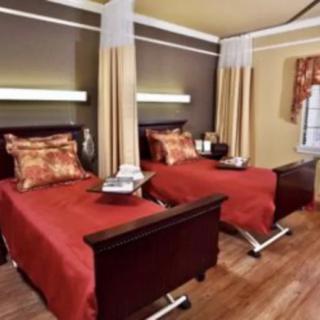 A hospital room with two neatly made twin beds, separated by curtains, featuring a warm color scheme and wooden flooring.