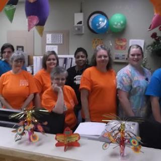 A group of staff members in orange shirts gathered at the office for a celebration.