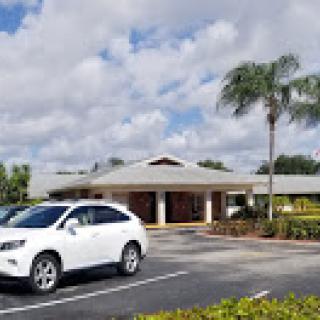 A modern building with palm trees in the foreground and a white car parked in the lot under a cloudy sky.