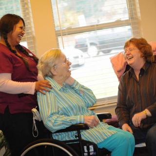 A caregiver and a visitor share a joyful moment with an elderly woman in a wheelchair.