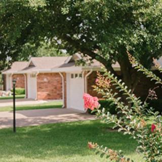 A suburban residential scene with a brick house, driveway, and lush greenery.