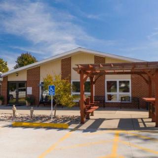 A small building with an entrance, flanked by a wooden pergola and surrounded by a parking area under a clear blue sky.