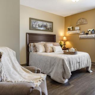 A cozy, neutral-toned bedroom with a neatly made bed, decorative shelves, and a comfortable chair.