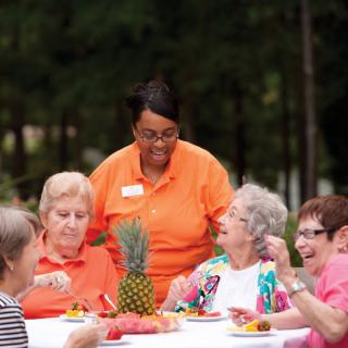 A group of seniors enjoying a meal together outdoors with a caregiver assisting them.