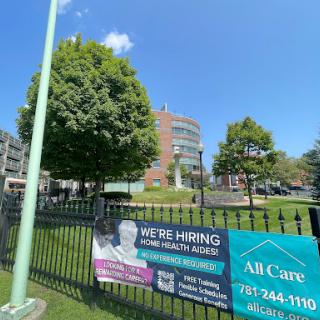 A view of a healthcare facility with a hiring sign in front, surrounded by greenery.