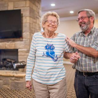 An elderly woman receiving support from a man indoors, both smiling happily.