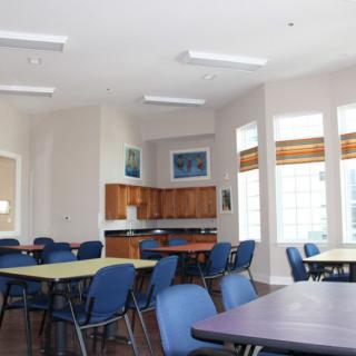An empty classroom with multiple tables and chairs, well-lit by large windows.