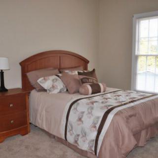 A neatly arranged bedroom with a bed featuring brown and beige bedding, a wooden headboard, a nightstand with a lamp, and a window with white trim.