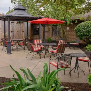 A cozy outdoor patio area featuring tables, chairs, a gazebo, and a red umbrella surrounded by lush greenery.
