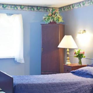 A cozy bedroom with blue walls, featuring a bed, a bedside table with a lamp, and a wardrobe.