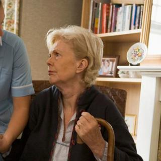 An elderly woman seated in a living room being assisted by a nurse.