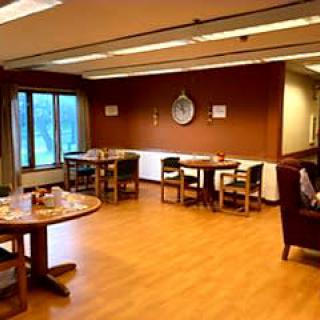 A cozy dining area with several tables and chairs, wooden flooring, and a large window providing natural light.