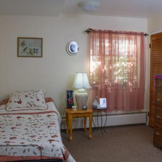 A cozy bedroom with a single bed, a lamp on a wooden table, and sunlight streaming through the curtains.