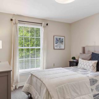 A cozy and well-lit bedroom featuring neutral tones, with a bed, lamp, window with curtains, and decorative artwork.