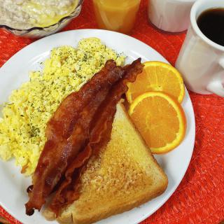 A classic breakfast with scrambled eggs, crispy bacon, toast, orange slices, and a cup of coffee.