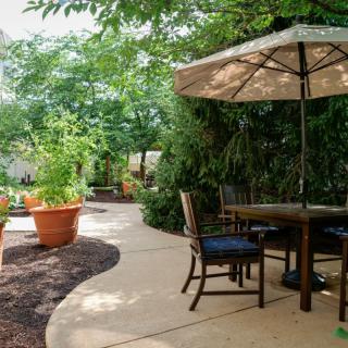 A peaceful garden patio with a table, chairs, and an umbrella surrounded by lush greenery and potted plants.