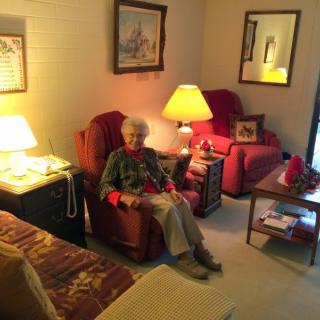 A cozy living room with an elderly person sitting in a chair, surrounded by warm lighting and comfortable furniture.