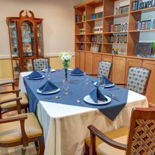 An elegantly set dining table surrounded by wooden bookshelves, featuring a blue and white tablecloth with folded napkins and a floral centerpiece.